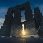 Low-angle view of the massive Chronicles of Georgia monument in Tbilisi, with the sun setting or rising through one of its arches, casting a bright glow.