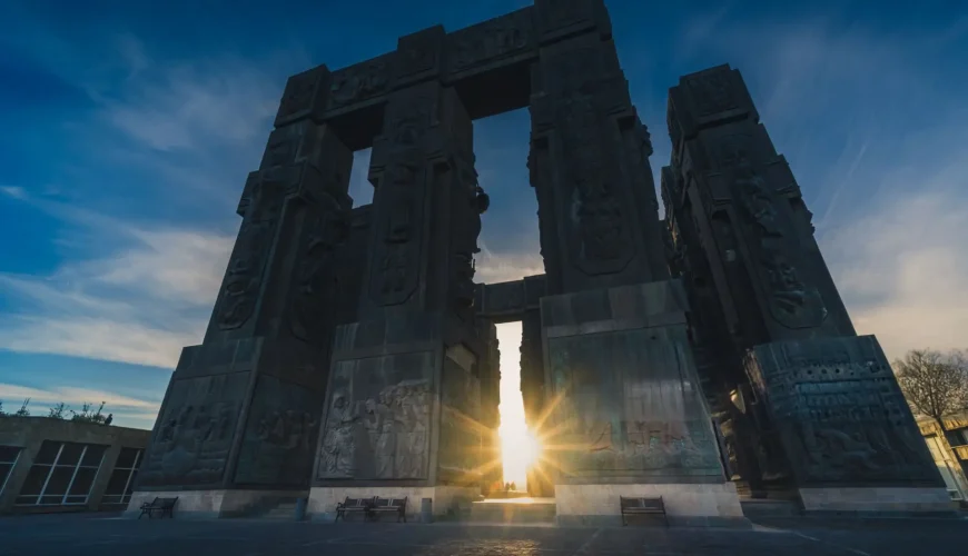 Low-angle view of the massive Chronicles of Georgia monument in Tbilisi, with the sun setting or rising through one of its arches, casting a bright glow.