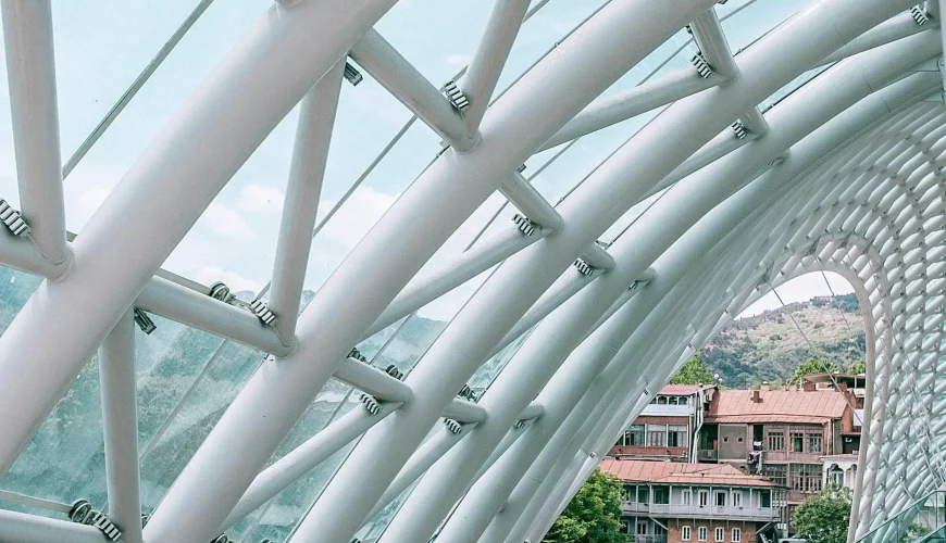 Close-up of the futuristic white steel and glass structure of the Bridge of Peace in Tbilisi, with a view of old town buildings and the Mtkvari River below.