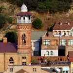 Close-up of the brick minaret of the Jumah Mosque in Tbilisi, with a unique octagonal cupola and arched windows, set against a backdrop of houses on a hillside.
