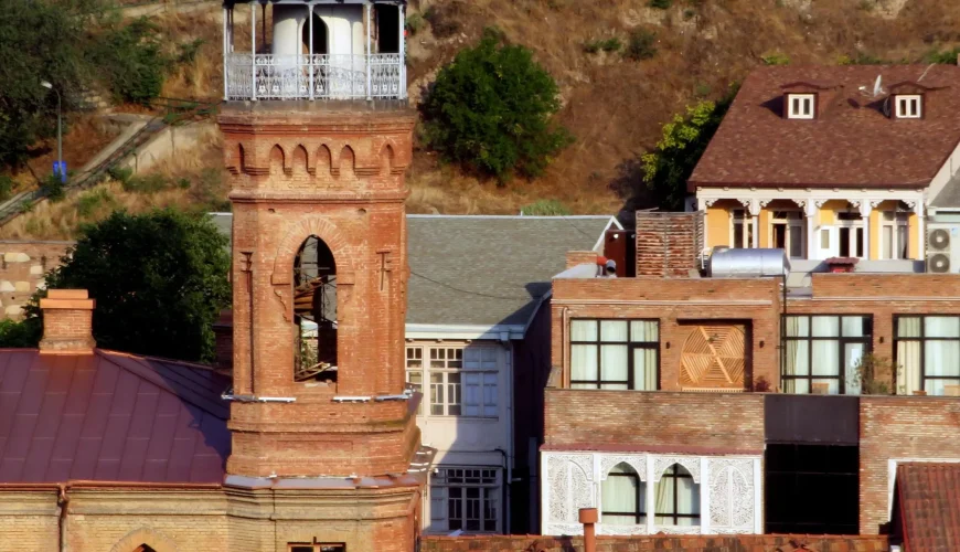 Close-up of the brick minaret of the Jumah Mosque in Tbilisi, with a unique octagonal cupola and arched windows, set against a backdrop of houses on a hillside.