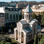 An aerial view of Kashueti St. George Church in Tbilisi, Georgia, surrounded by trees and modern buildings along Rustaveli Avenue.