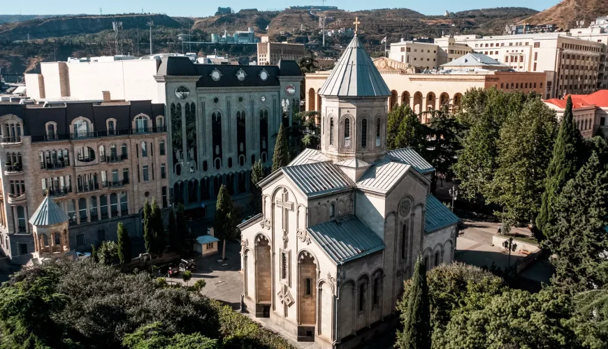 An aerial view of Kashueti St. George Church in Tbilisi, Georgia, surrounded by trees and modern buildings along Rustaveli Avenue.