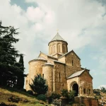 A low-angle view of the historic Metekhi Church in Tbilisi, Georgia, with its stone dome and a cross on top, framed by tall cypress trees against a cloudy sky.