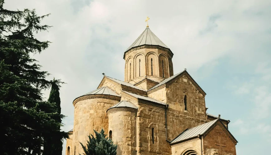 A low-angle view of the historic Metekhi Church in Tbilisi, Georgia, with its stone dome and a cross on top, framed by tall cypress trees against a cloudy sky.