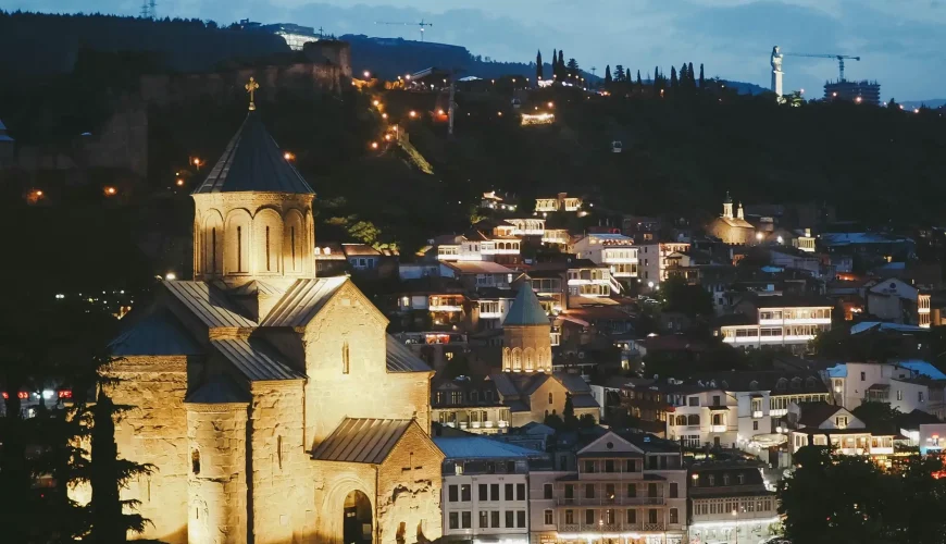 A beautifully lit Metekhi Church at night, with Tbilisi's Old Town sparkling on the hillside behind it.
