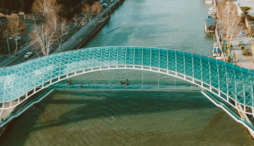 An aerial view of the Bridge of Peace in Tbilisi, Georgia, with the Kura River flowing underneath and a mix of historic and modern city architecture visible on both banks
