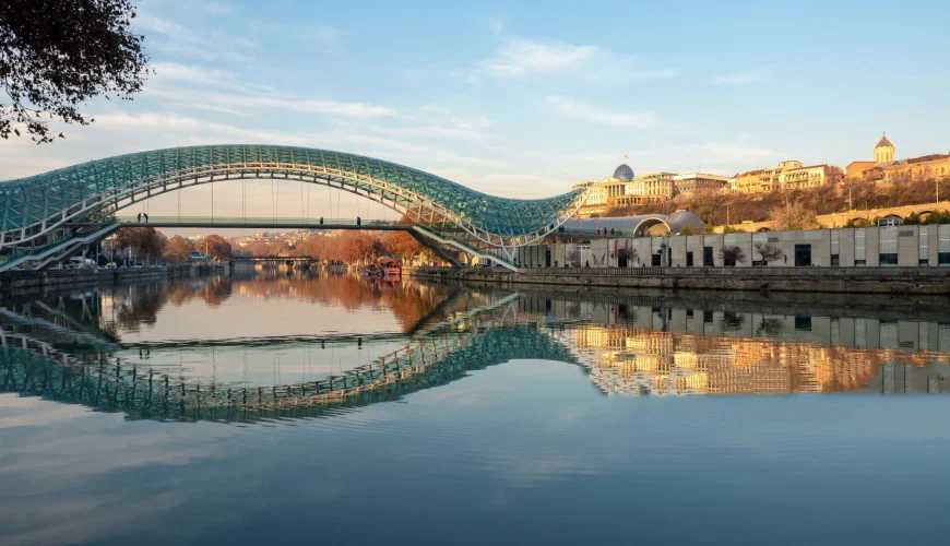 The Bridge of Peace in Tbilisi, Georgia, reflected in the calm waters of the Kura River at sunset, with the Old Town visible in the background.