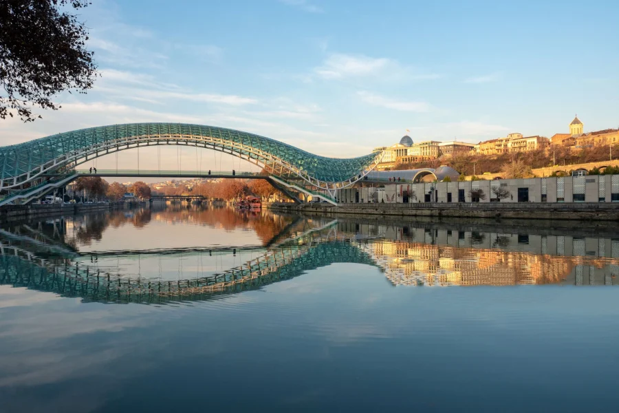 The Bridge of Peace in Tbilisi, Georgia, reflected in the calm waters of the Kura River at sunset, with the Old Town visible in the background.