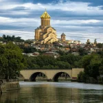 Panoramic view of the Holy Trinity Cathedral (Sameba) in Tbilisi, Georgia, with a stone bridge and the Mtkvari River in the foreground.