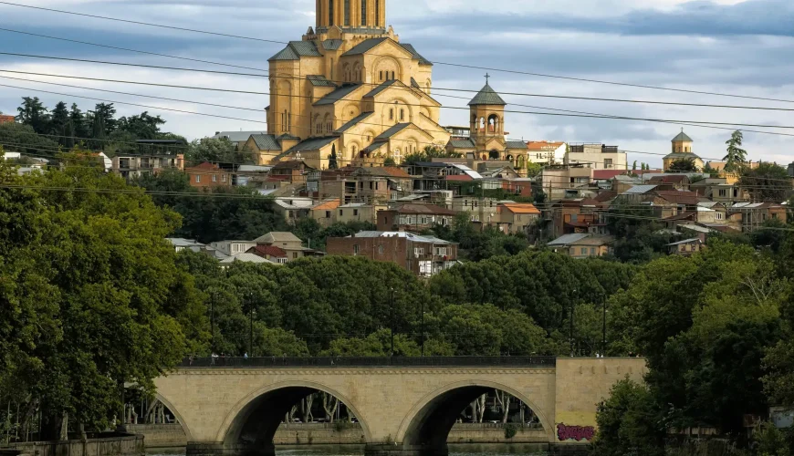 Panoramic view of the Holy Trinity Cathedral (Sameba) in Tbilisi, Georgia, with a stone bridge and the Mtkvari River in the foreground.