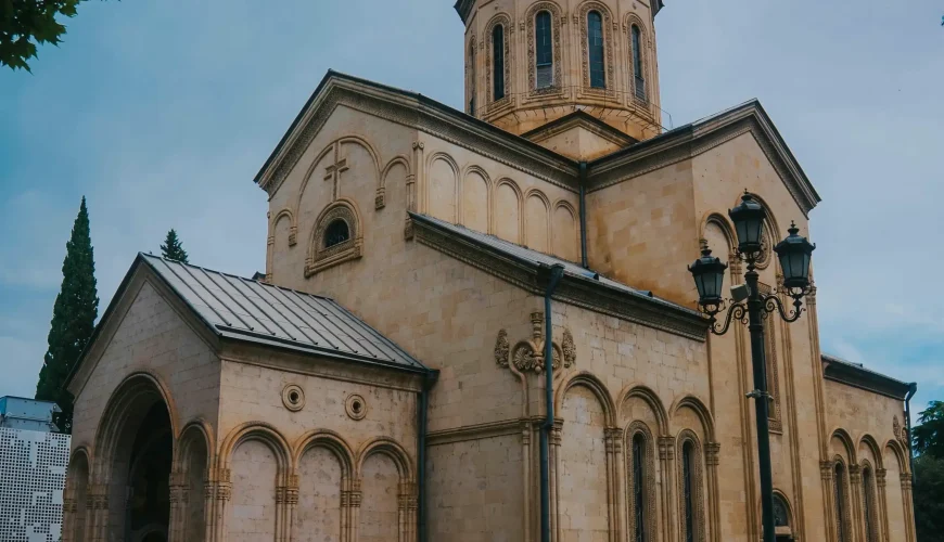 The Sioni Cathedral of the Dormition in Tbilisi, Georgia, featuring its stone facade, prominent dome, and arched windows under a cloudy sky.