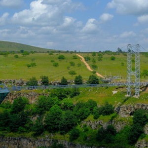 A panoramic view of the Tsalka Canyon Diamond Bridge, showing the suspended walkway and the diamond-shaped observation deck against a backdrop of rolling green hills and a cloudy sky.