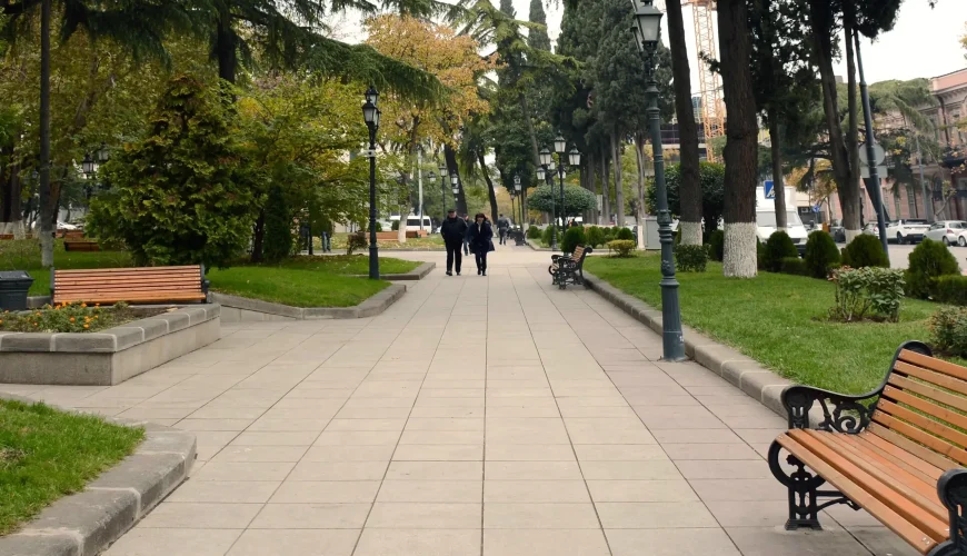 A long, paved pathway in April 9 Park, Tbilisi, lined with tall evergreen trees, park benches, and distant figures walking.