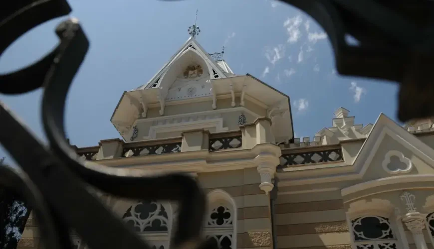 A detailed view of the ornate upper facade and dome of the Art Palace of Georgia (Museum of Cultural History) in Tbilisi, seen through a decorative metal gate, under a blue sky.
