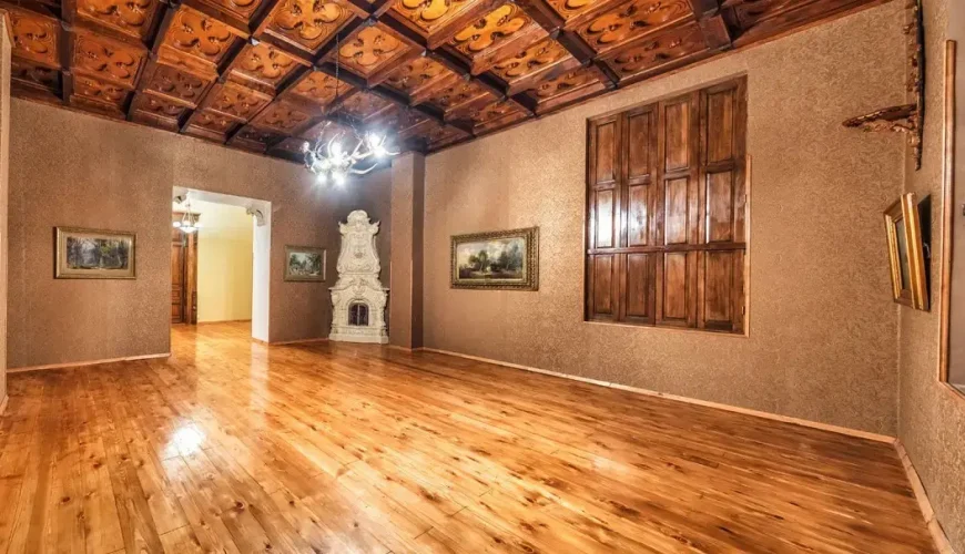 A grand, empty room inside the Art Palace of Georgia (Museum of Cultural History) in Tbilisi, featuring an intricately carved wooden ceiling, an ornate white fireplace, and polished wooden floors