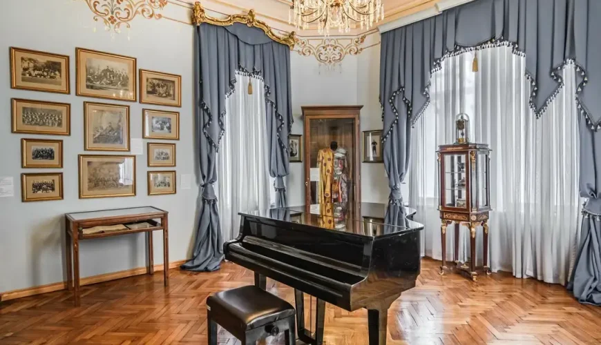 A beautifully decorated room at the Art Palace of Georgia (Museum of Cultural History) in Tbilisi, featuring a black grand piano, elaborate blue and white drapes, and a wall adorned with framed historical photos and portraits.