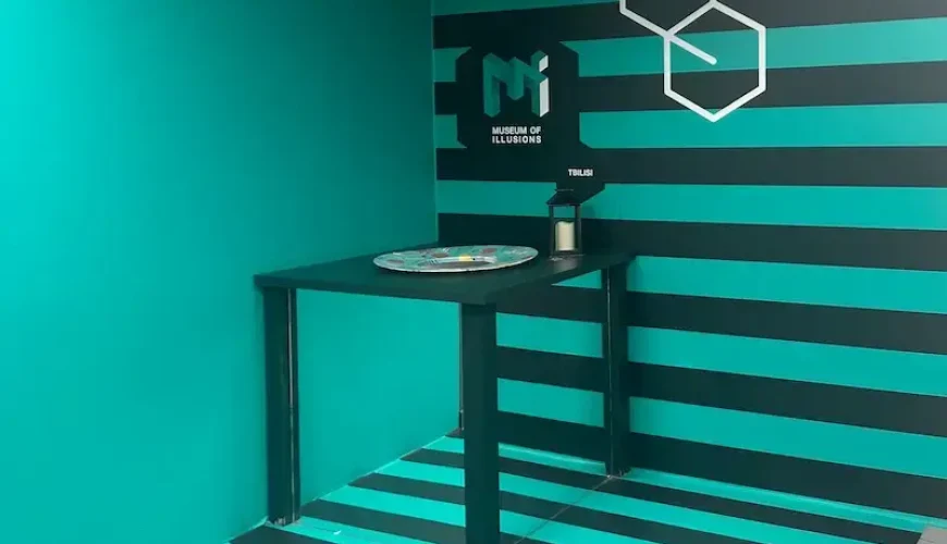 An optical illusion room at the Museum of Illusions in Tbilisi, featuring a corner setup with a table, painted in teal and black stripes to create a distorted perspective.