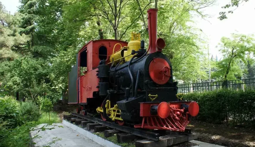The station building of the World's First Children's Railway in Mushtaidi Park, Tbilisi, with a small red and white locomotive and railway tracks visible