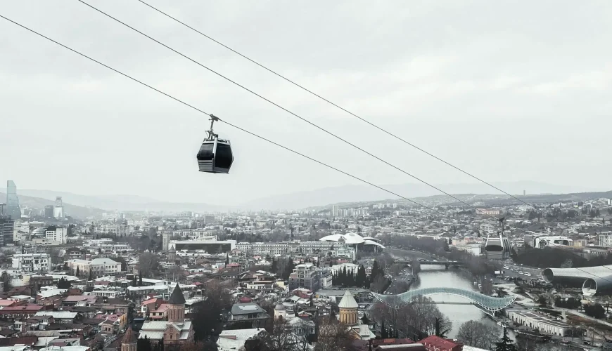 Aerial view of Tbilisi city with a cable car cabin suspended over rooftops, showing the Mtkvari River, bridges, and distant mountains under an overcast sky.