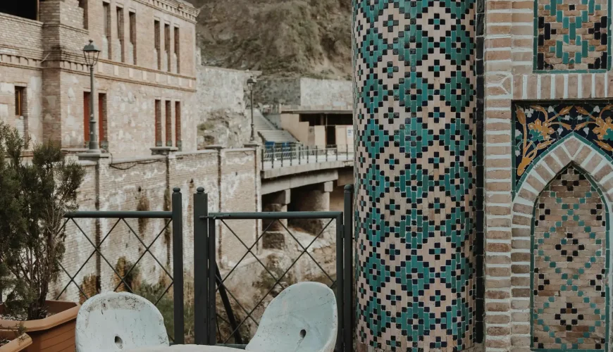 A patio area with a stone table and chairs next to an ornate tiled column and brick wall in Tbilisi's historic Abanotubani district.