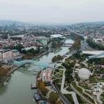 Panoramic aerial view of Tbilisi, Georgia, showing the Bridge of Peace, Rike Park, the Kura River, and the cityscape under a cloudy sky.