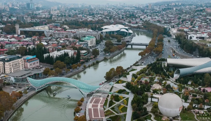 Panoramic aerial view of Tbilisi, Georgia, showing the Bridge of Peace, Rike Park, the Kura River, and the cityscape under a cloudy sky.