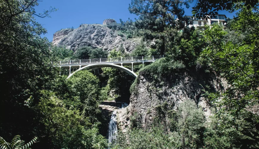 Bridge over a waterfall in the lush Tbilisi Botanical Garden, surrounded by green trees and rocky cliffs under a clear blue sky