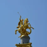 Close-up of the golden statue of St. George slaying a dragon, atop a tall monument in Freedom Square, Tbilisi, against a clear blue sky.