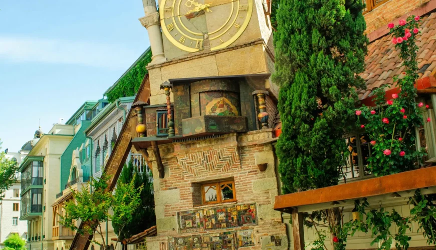 The leaning clock tower of the Rezo Gabriadze Marionette Theater in Tbilisi, with a detailed, eclectic design, surrounded by a narrow street and buildings.