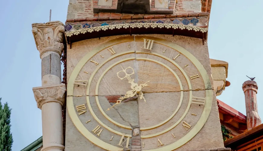 A close-up of the large, detailed clock face and eclectic architectural elements of the leaning Gabriadze Marionette Theater clock tower in Tbilisi.