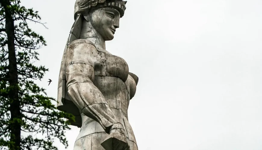 Side profile of the Kartlis Deda (Mother of Georgia) statue in Tbilisi, showing her from a low angle against a cloudy sky, with a tall tree in the foreground.