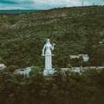 The Kartlis Deda (Mother of Georgia) statue standing on a hill overlooking Tbilisi, holding a sword and a bowl, surrounded by a lush forest.
