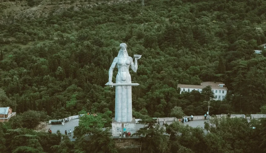 The Kartlis Deda (Mother of Georgia) statue standing on a hill overlooking Tbilisi, holding a sword and a bowl, surrounded by a lush forest.