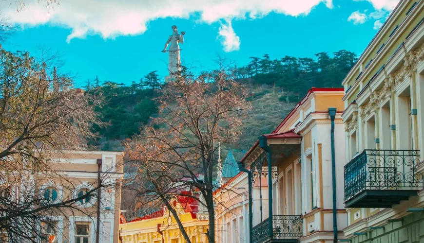 Kartlis Deda (Mother of Georgia) statue visible on a hill above colorful traditional buildings and a street in Tbilisi Old Town.