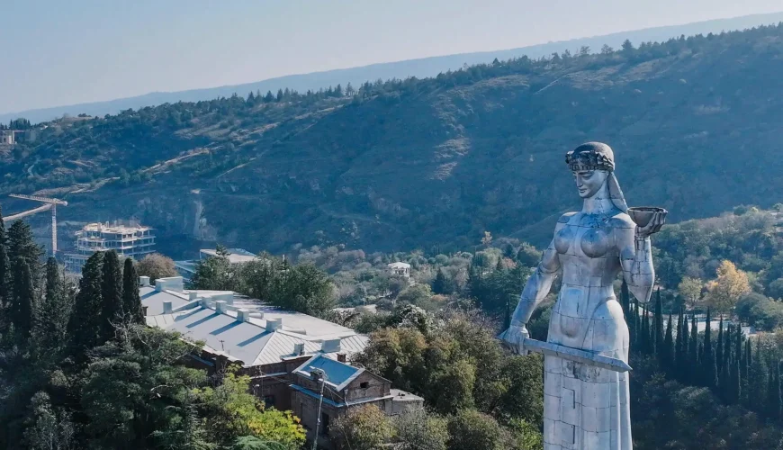 The Kartlis Deda (Mother of Georgia) statue in Tbilisi, Georgia, with a panoramic view of the forested hills and city buildings in the background.