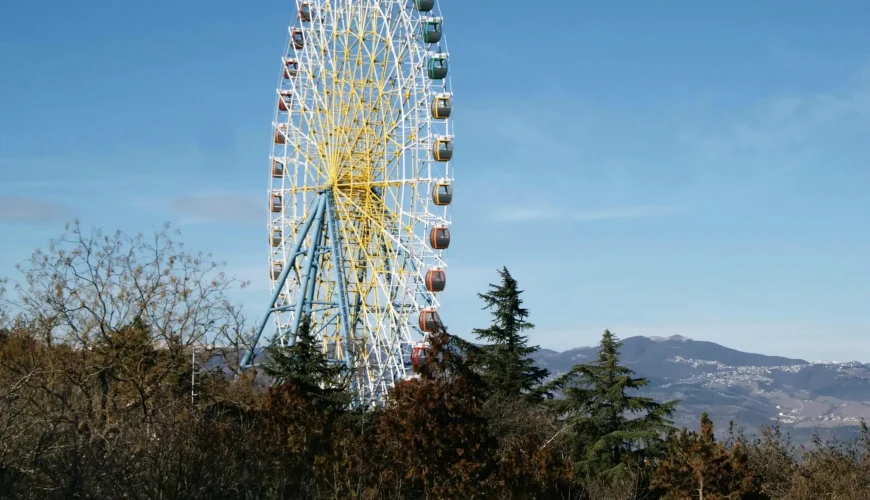 The towering Ferris Wheel at Mtatsminda Park in Tbilisi, Georgia, visible above treetops with a clear blue sky and distant mountains.