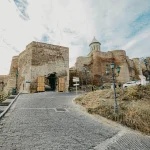 Stone entrance gate to Narikala Fortress in Tbilisi, Georgia, with a cobblestone path leading up and an old church visible on the fortress wall