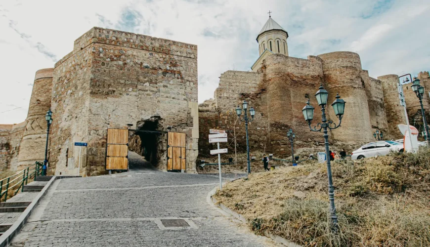 Stone entrance gate to Narikala Fortress in Tbilisi, Georgia, with a cobblestone path leading up and an old church visible on the fortress wall