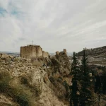 Panoramic view of the ancient stone ruins of Narikala Fortress overlooking Tbilisi city, with rocky terrain and trees in the foreground.