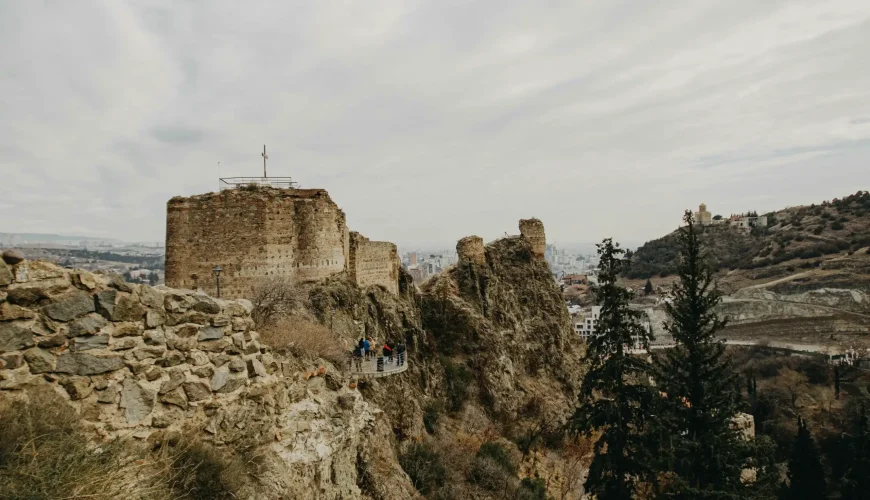 Panoramic view of the ancient stone ruins of Narikala Fortress overlooking Tbilisi city, with rocky terrain and trees in the foreground.
