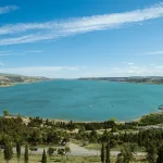 Panoramic view of Turtle Lake (Kus Tba) in Tbilisi, Georgia, showing the expansive turquoise lake, surrounding green hills, a small beach, and distant city structures under a blue sky.