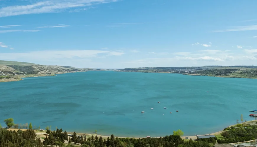 Panoramic view of Turtle Lake (Kus Tba) in Tbilisi, Georgia, showing the expansive turquoise lake, surrounding green hills, a small beach, and distant city structures under a blue sky.