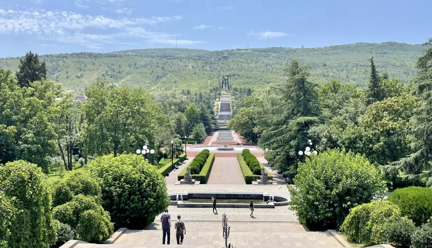 Panoramic view of Vake Park in Tbilisi, Georgia, showing a vast terraced staircase, water features, manicured gardens, and distant green hills under a blue sky.