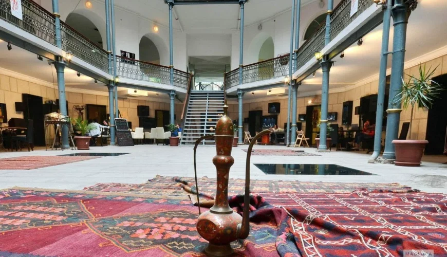 The grand multi-story interior of Carvasla – Tbilisi Museum, featuring a central open space with patterned rugs, an antique copper vessel, and a visible staircase leading to upper levels, showcasing its historic architecture.