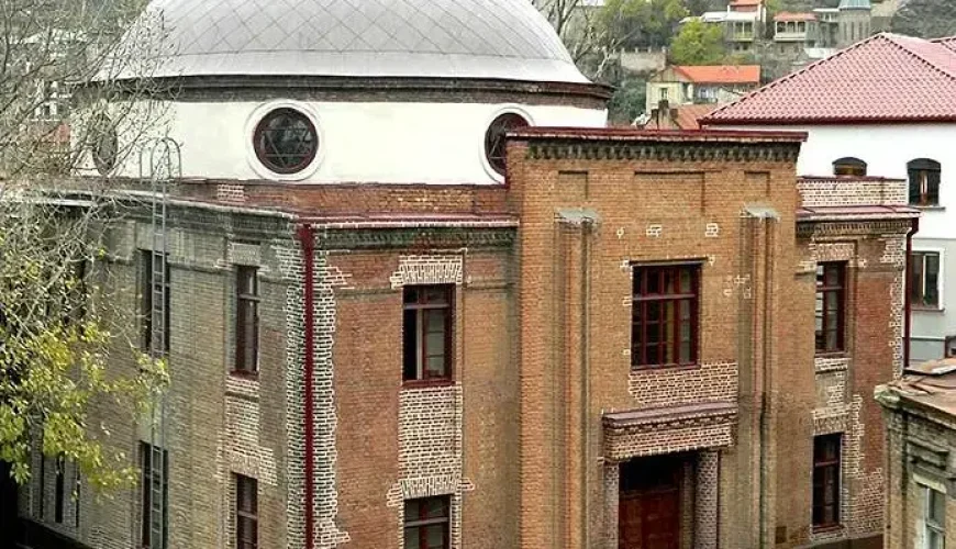 The historic brick exterior of the David Baazov Museum of History of Jews of Georgia in Tbilisi, featuring a prominent domed roof and traditional architectural details, set amidst other city buildings.