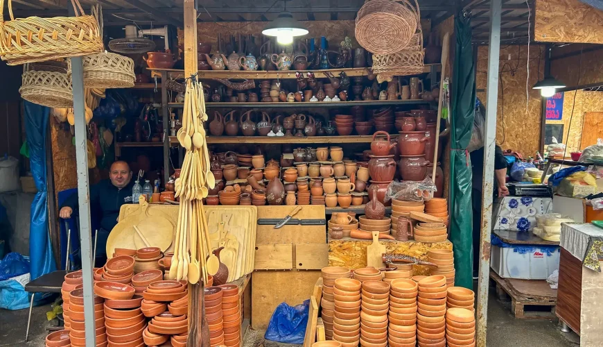 A vendor sitting in front of a stall overflowing with handmade clay pottery, baskets, and wooden spoons at Dezerteer Bazaar in Tbilisi