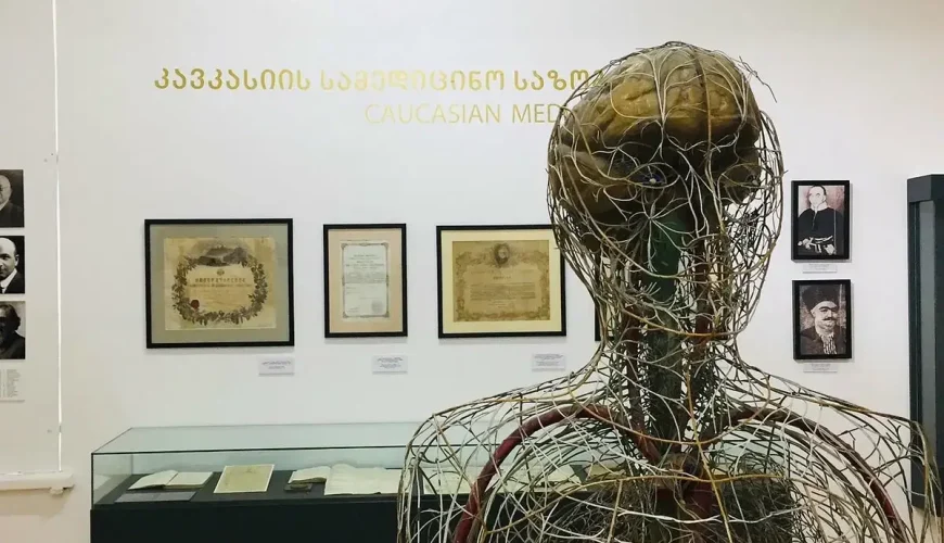 An exhibition hall inside the Georgian Medicine History Museum in Tbilisi, featuring a large wire anatomical model of a human body, framed historical documents, and portraits, with 'CAUCASIAN MEDICINE' on the wall.