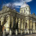 The stately yellow and white facade of the Georgian Medicine History Museum in Tbilisi, Georgia, featuring a prominent dome and classical architectural details, framed by bare trees.