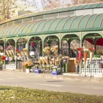 A wide view of the Orbeliani Flower Market in Tbilisi, with vendors selling flowers under a long, green-domed structure on a sunny day